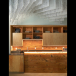 Exterior view of Hugo House, showcasing a contemporary architectural design with a striking facade that features large windows and a mix of materials, including wood and metal. The building is set in an urban environment, surrounded by greenery and pedestrian pathways. The design emphasizes openness and creativity, reflecting its role as a community space for writers and literary events.