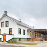 Exterior view of Fort Worden Building 305, showcasing a historic structure with a charming architectural style. The building features a combination of wood and brick materials, large windows, and a gabled roof. Surrounded by lush greenery and open space, the setting highlights the building's integration with the natural landscape, reflecting its significance as a community space.