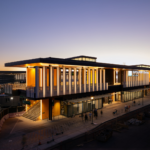 Exterior view of the Seattle Ferry Terminal, featuring a modern architectural design with a large, open structure and a prominent roofline. The terminal is bustling with activity, showcasing ferries docked at the waterfront, passengers boarding, and a backdrop of the Seattle skyline and waterfront. The scene captures the vibrant atmosphere of the terminal as a key transportation hub.