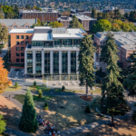 The exterior of the Founders Hall at the Foster School of Business, featuring a modern architectural design with large glass windows and a sleek facade. The building is surrounded by landscaped greenery and pathways, set against a clear sky.