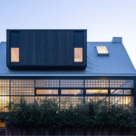 A modern residential home with a sleek design, featuring large windows and a combination of wood and stone materials. The house is surrounded by greenery, showcasing a well-maintained lawn and landscaping.
