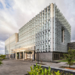 The exterior of the new U.S. Embassy in Guatemala, showcasing a modern architectural design with a prominent entrance, large glass windows, and a landscaped courtyard. The building features a blend of stone and metal materials, set against a clear blue sky.