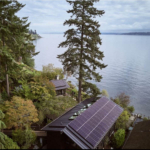 The image depicts the Loom House, a meticulously renovated 1960s mid-century modern home located on Bainbridge Island, Washington. The residence showcases clean lines, expansive windows, and natural wood finishes, harmoniously blending with the surrounding lush Pacific Northwest landscape. Overlooking Puget Sound, the property features sustainable design elements, including solar panels on the roof and rainwater collection systems, reflecting its Living Building Challenge 4.0 Certification.