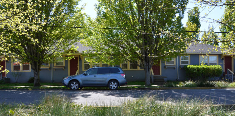 The image shows a neighborhood street with a four-plex building. There is a car in front and it is a tree-lined street.